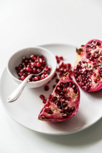 Aufgeschnittener roter Granatapfel auf weißem Teller und weißem Hintergrund, lose Kerne in weißer Schale mit Silberlöffel Aufgeschnittener roter Granatapfel auf weißem Teller und weißem Hintergrund, lose Kerne in weißer Schale mit Silberlöffel